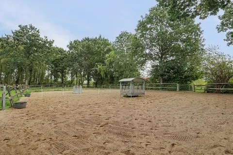 Zandbak van de rijbak van 20 bij 40 meter met overdekte hooiruif, omzoomd door bomen